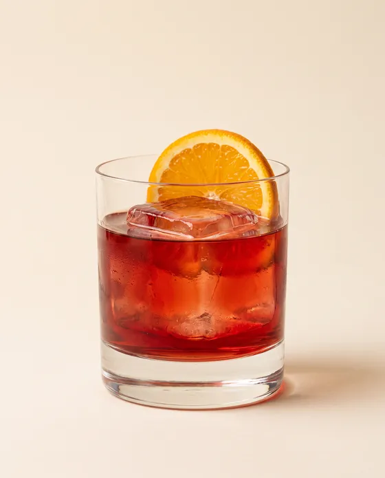 Negroni — a rocks glass with a red-amber cocktail over a large ice cube and an orange peel, on a cream background