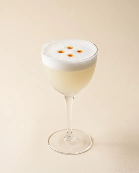 Pisco Sour — a glass with a white-foam sour crowned by three drops of Angostura, on a cream background