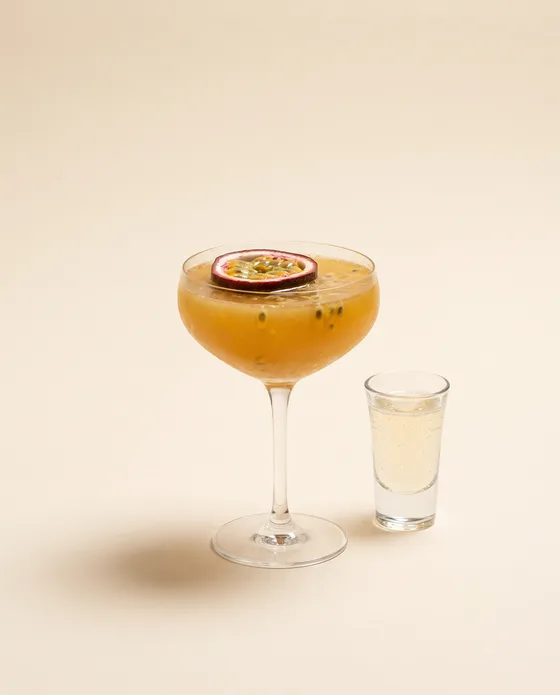 Pornstar Martini — a coupe with a golden passion-fruit cocktail, half a passion fruit floating and a cava shot on the side, on a cream background